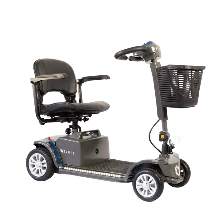 The Reyhee Cruiser Portable Electric Mobility Scooter is compact and four-wheeled, featuring a foldable tiller for easy storage, a cushioned seat with armrests, and a steering handle with an attached basket. It comes in gray with white-rimmed wheels and a small front light.