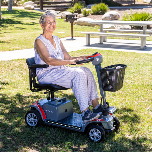 An elderly woman beams as she navigates the park on her Reyhee Cruiser Portable Electric Mobility Scooter with Front Basket, wearing a striped sleeveless outfit. Little treasures fill the basket while a picnic table and grill invite nearby amidst lush grass and trees.