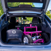 The car trunk opens to reveal a Reyhee EXO Portable 4-Wheel Mobility Scooter with a pink frame, nestled alongside a Reyhee bag. Green trees and a sunny outdoor setting serve as the backdrop.