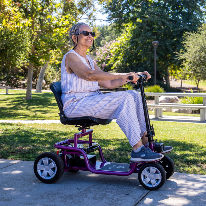 Wearing sunglasses and a striped sleeveless outfit, an individual rides a purple Reyhee EXO Portable 4-Wheel Mobility Scooter along a sunny park path, with green grass, trees, and benches in the backdrop.