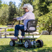 A smiling elderly woman with white hair enjoys her Reyhee EXO Portable 4-Wheel Mobility Scooter in a park. Wearing a light shirt, jeans, and a sun hat, she glides effortlessly on grass amidst trees and benches, experiencing freedom and joy with every ride.