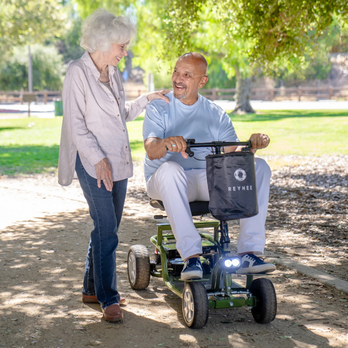 An elderly woman with white hair smiles, walking beside a man on his Reyhee EXO Portable 4-Wheel Mobility Scooter. They enjoy a sunny day in the park, appearing happy amidst green trees and natural light.