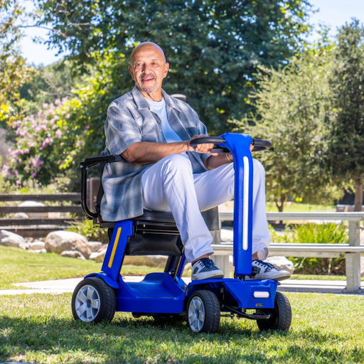 A man smiles on a Reyhee Flex Electric Folding Mobility Scooter in a park, highlighting its foldable design. Dressed in a plaid shirt, white pants, and sneakers, he enjoys the sunny day surrounded by trees and a wooden fence.