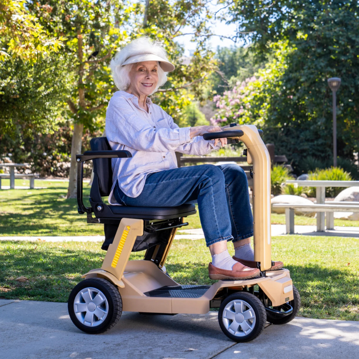 An elderly woman in a white hat and light blue shirt smiles while sitting on a beige Reyhee Flex Electric Folding Mobility Scooter in a park, surrounded by greenery and trees as sunlight filters through the leaves.