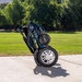 The Reyhee Roamer Folding Long Range Electric Wheelchair, which is FDA-cleared and equipped with dual brushless DC motors, stands upright on a concrete path in a park. In the background are a lush green lawn and trees under a clear blue sky.