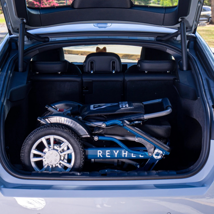 A Reyhee Roamer Folding Long Range Electric Wheelchair with dual brushless DC motors is folded compactly in an open car trunk, fitting neatly into the space while revealing the cars interior and part of the exterior environment.