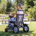 An elderly person with short grey hair sits in a Reyhee Roamer Folding Long Range Electric Wheelchair on a sunny, grassy area. Wearing a light-colored dress, they face away with trees and a wooden picnic table behind them, effortlessly powered by dual brushless DC motors.