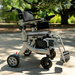 The Reyhee Superlite 3-in-1 Electric Foldable Wheelchair with joystick control sits on a sunlit pathway, surrounded by green trees and a wooden fence, suggesting a park setting that highlights its lightweight, travel-friendly design and versatile mobility solutions.