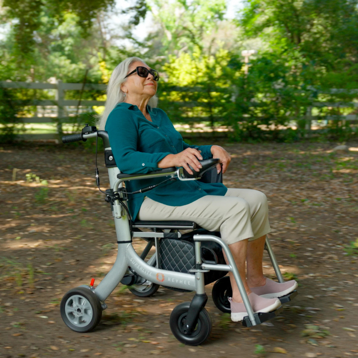 A woman in a teal shirt and beige pants, wearing sunglasses, smiles as she sits in the Reyhee Superlite 3-in-1 Electric Foldable Wheelchair. The sleek, lightweight design complements the sunny outdoor setting with trees and a wooden fence in the background.