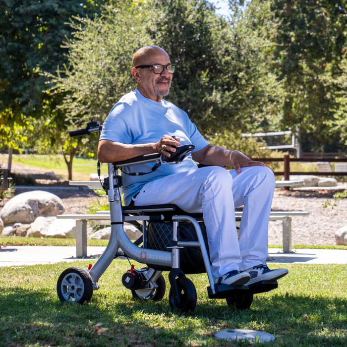 A man in a light blue shirt and white pants smiles as he navigates the sunny park in his Reyhee Superlite 3-in-1 Electric Foldable Wheelchair. Trees and boulders form a serene background, enhancing his joyful journey.