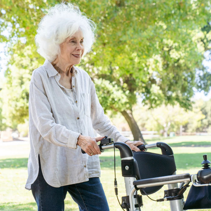 An elderly woman with white hair uses a Reyhee Superlite 3-in-1 Electric Foldable Wheelchair in a sunny park. She wears a light beige shirt and dark pants, smiling with green trees and grass in the background.