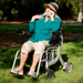 An elderly woman in a blue shirt and beige pants sits in a Reyhee Superlite 3-in-1 Electric Foldable Wheelchair on the grass. Shes wearing a wide-brimmed hat and smiling while adjusting her sunglasses, as sunlight filters through the trees, highlighting her versatile mobility solution.