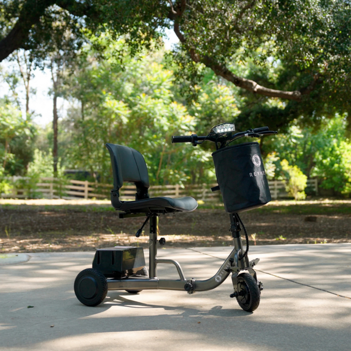 The Reyhee Triad Compact Folding Electric Mobility Scooter, lightweight and portable with three wheels, a black seat, and front basket, is parked on a sunlit path in the park. It complements the lush greenery and trees beautifully, creating a serene outdoor setting.