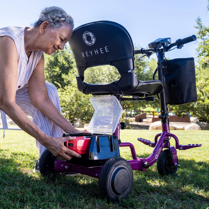 An elderly person with short gray hair places a red tool in the storage compartment of a Reyhee Triad Compact Folding Electric Mobility Scooter, which is purple and lightweight. The scooter, featuring a black seat with REYHEE on it, sits on the grass amid trees and sunlight.