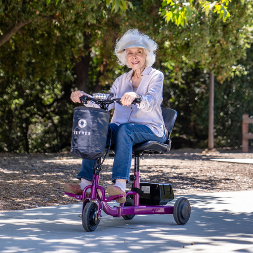 An elderly woman with white hair smiles while riding a Reyhee Triad Compact Folding Electric Mobility Scooter in purple. She wears a light blouse, jeans, and slippers as she enjoys the parks paved path surrounded by trees.