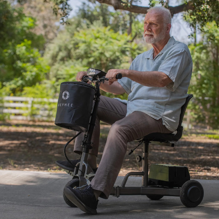 An older man with a white beard rides a Reyhee Triad Compact Folding Electric Mobility Scooter with a Swap & Go Battery System on a parks paved path. Hes wearing a light blue shirt and brown pants, surrounded by trees and greenery. A branded bag is attached to the scooter.