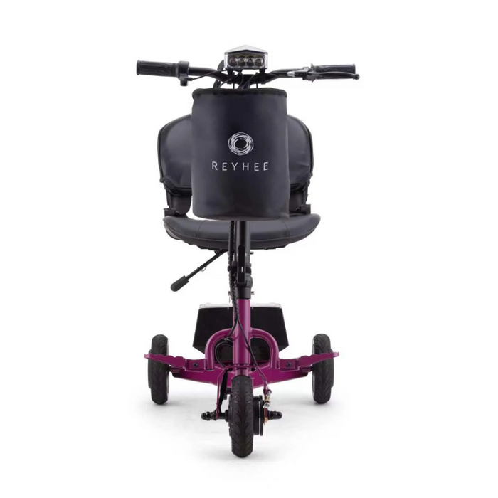 The REYHEE Triad Compact Folding Electric Mobility Scooter boasts a pink frame, three wheels, a cushioned seat, small headlights above a front storage bag, and the Swap & Go Battery System. Its handlebars with grips enhance both practicality and charm.