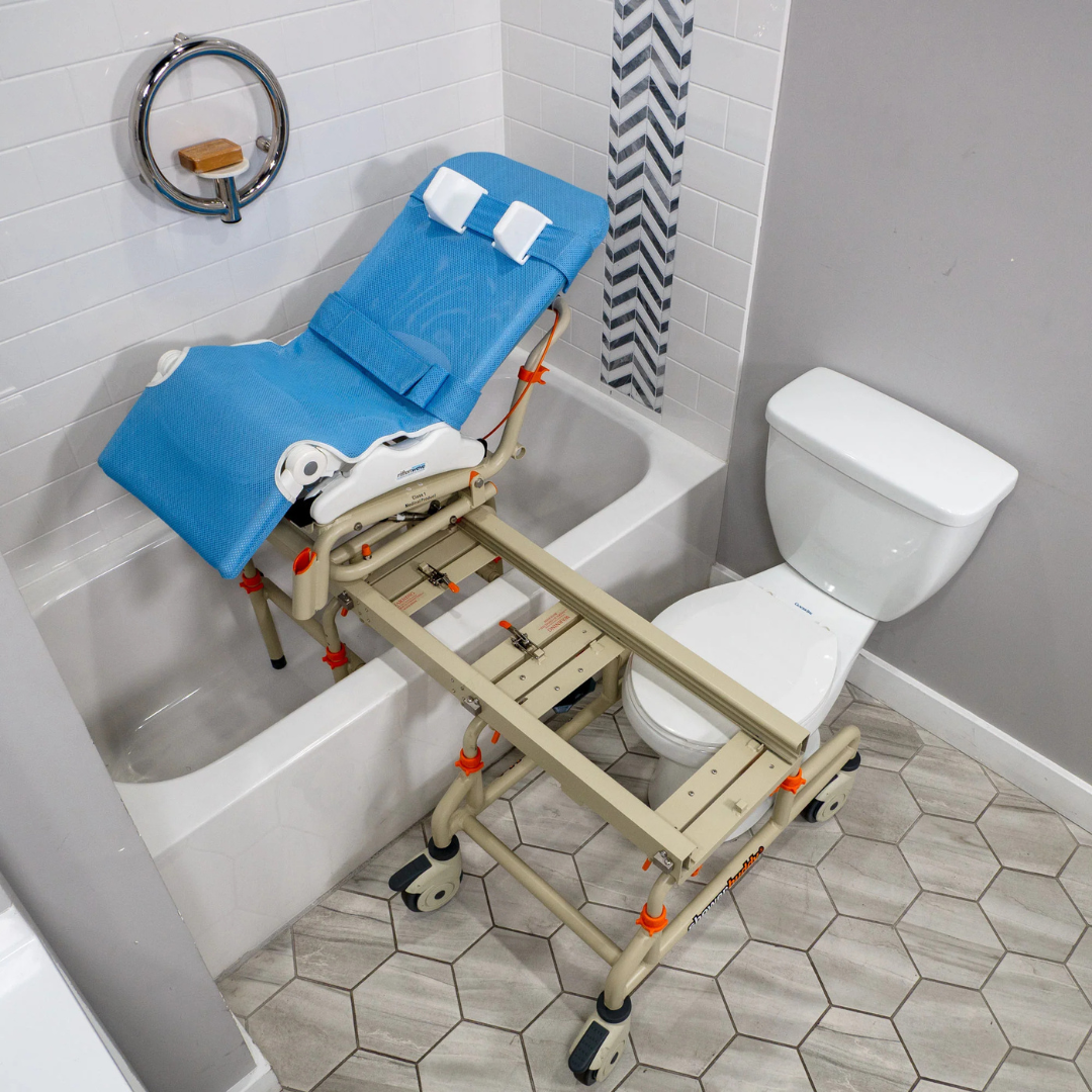 A beige and blue Rifton Wave Pediatric Shower Chair with an adjustable frame and sliding rail system is placed over a bathtub and toilet in a bathroom featuring hexagonal floor tiles and white tiled walls.