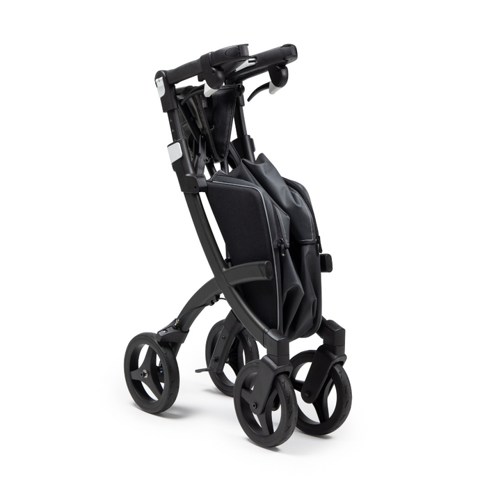 The folded Rollz FLEX 2 Lightweight Rollator Walker in black stands upright, showcasing its compact design with four wheels, a sturdy frame, and a comfortable handlebar for portability and ease of use. It also features an XL storage bag and seat for on-the-go convenience.