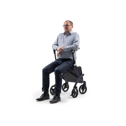 A man in glasses and a blue shirt is seated, facing forward, on a Rollz FLEX 2 Lightweight Rollator Walker featuring four wheels, a compact design, and an XL storage bag. The white background highlights this mobility aid.