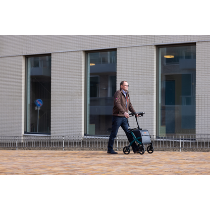 A man in a brown jacket and jeans strolls outdoors on a paved path next to a modern building with large windows, enjoying the quiet surroundings while using the Rollz FLEX 2 Lightweight Rollator Walker equipped with an XL storage bag for essentials.