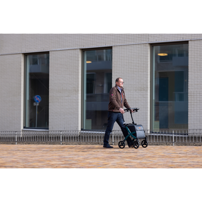 A man in a brown jacket and jeans strolls outdoors on a paved path next to a modern building with large windows, enjoying the quiet surroundings while using the Rollz FLEX 2 Lightweight Rollator Walker equipped with an XL storage bag for essentials.