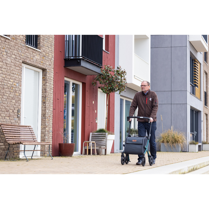 A man strolls with his Rollz FLEX 2 Lightweight Rollator Walker with XL Storage Bag & Seat along a modern residential building featuring colorful facades. Nearby, a wooden bench and potted plants by the entrance create a calm suburban vibe.