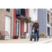 A man strolls with his Rollz FLEX 2 Lightweight Rollator Walker with XL Storage Bag & Seat along a modern residential building featuring colorful facades. Nearby, a wooden bench and potted plants by the entrance create a calm suburban vibe.