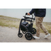 A person in a navy jacket and beige pants walks on a gravel path using the Rollz Motion Electric 3-in-1 Rollator, Transport Chair & Powerchair next to grass by water. This multifunctional aid features sturdy wheels and has a storage compartment for essentials.