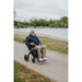 A smiling older man in a blue jacket and beige pants sits happily in a Rollz Motion Electric 3-in-1 Rollator, Transport Chair & Powerchair by a lakeside path, wearing glasses and surrounded by grassy areas and trees beneath a cloudy sky.