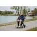 A bearded man in a navy jacket and beige pants smiles while using his Rollz Motion Electric 3-in-1 Rollator, Transport Chair & Powerchair along a gravel path by a calm lake, trees and a house in the background enhancing his journey.