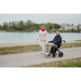 An elderly couple enjoys a lakeside stroll. The man sits comfortably in a Rollz Motion Electric 3-in-1 Rollator, Transport Chair & Powerchair, wearing sunglasses and a dark jacket, while the woman accompanies him in a light outfit and red scarf. Both look content as they travel along the path.