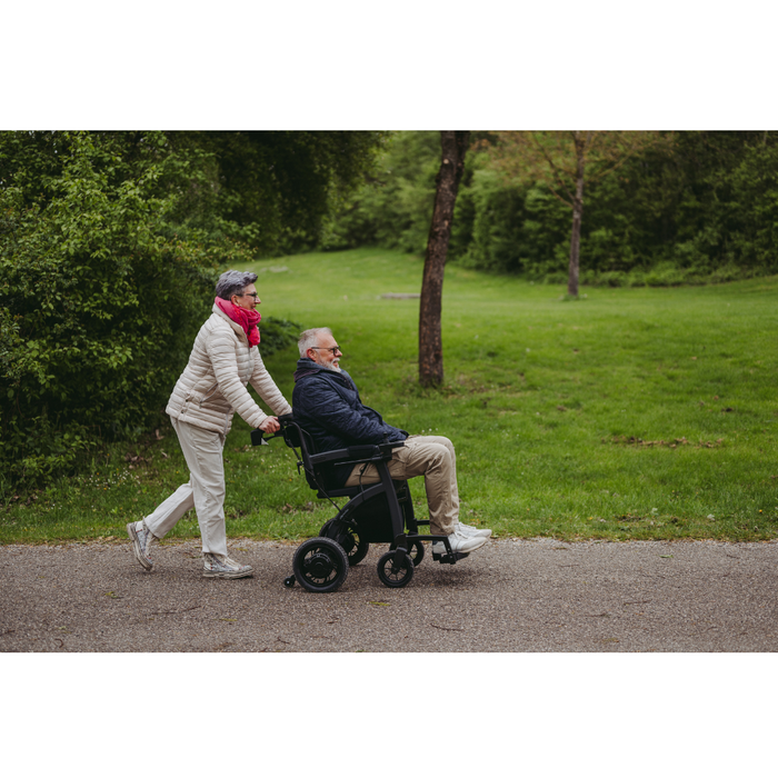 A senior is gently pushed by another elderly person in a Rollz Motion Electric 3-in-1 Rollator, Transport Chair, and Powerchair along a paved path through a lush park. Both are dressed warmly, enjoying the serene scene under an overcast sky with diffused lighting.