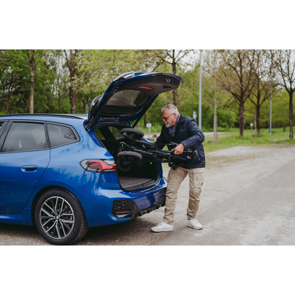 A man wearing a black jacket and beige pants loads a folded Rollz Motion Electric 3-in-1 Rollator, Transport Chair & Powerchair into the open trunk of a blue car while fresh green-leaved trees stand in the park background.