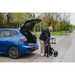 An elderly person stands beside an open blue car trunk on a tree-lined road under overcast skies, folding their black walker. This scene subtly highlights the convenience of the Rollz Motion Electric 3-in-1 Rollator, Transport Chair & Powerchair, blending comfort and versatility.