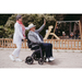 An older man in a Rollz Motion Electric 3-in-1 Rollator, Transport Chair & Powerchair waves happily as an older woman pushes him through the park. Both wear casual hats, with a playground and children in the background, creating a joyful scene for all.