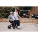 A man smiles from his Rollz Motion Electric 3-in-1 Rollator, Transport Chair & Powerchair at a woman in a white outfit with a pink scarf. Outdoors near a playground, kids and adults enjoy the freedom this mobility solution provides.