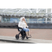 A person in a white coat with a fur-lined hood sits on the paved area in a Rollz Motion Electric 3-in-1 Rollator, Transport Chair & Powerchair, highlighting its practicality. They wear blue jeans and tan boots, with a modern building featuring metal structures in the background.