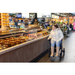 A woman using the Rollz Motion Electric 3-in-1 Rollator, Transport Chair & Powerchair, dressed in a white coat and boots, admires cheeses and sausages at a bustling indoor market filled with diverse food items and vibrant signs.