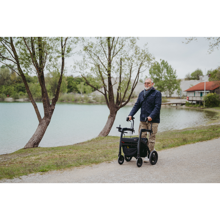 A bearded older man strolls along a lakeside path with his Rollz Motion Electric 3-in-1 Rollator, Transport Chair & Powerchair. Dressed in a blue jacket and beige pants, he walks past trees and calm water on an overcast day, with buildings in the distance.