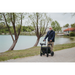 A bearded older man strolls along a lakeside path with his Rollz Motion Electric 3-in-1 Rollator, Transport Chair & Powerchair. Dressed in a blue jacket and beige pants, he walks past trees and calm water on an overcast day, with buildings in the distance.