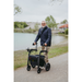 An elderly bearded man in a blue jacket and khaki pants navigates the lakeside path with the Rollz Motion Electric 3-in-1 Rollator, Transport Chair & Powerchair. Green trees and a red-roofed building under an overcast sky emphasize this modern mobility aids innovation.