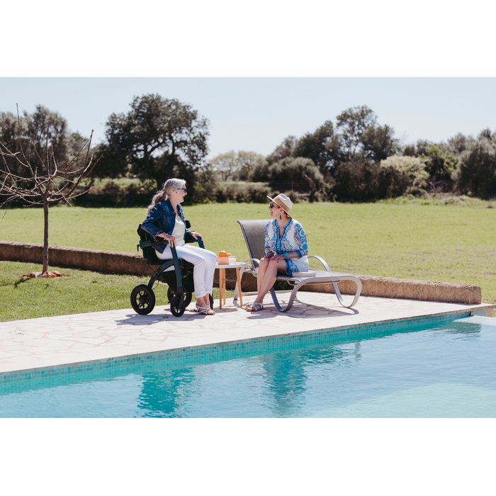 Two women lounge by the swimming pool on a sunny day. One sits comfortably in a Rollz Motion Performance All-Terrain 2-In-1 Rollator Transport Chair, while the other relaxes on a lounge chair. They chat animatedly, sipping drinks from a small table between them, surrounded by lush greenery.