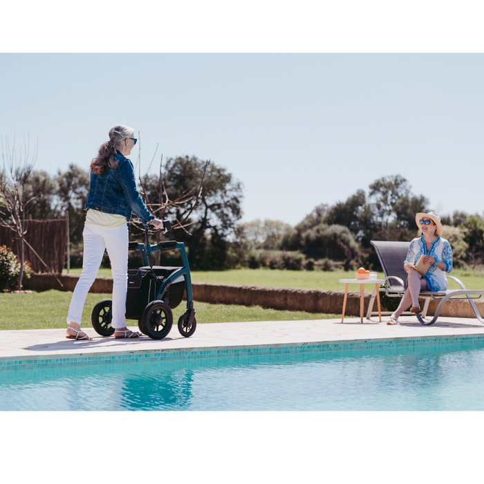 An elderly woman with a Rollz Motion Performance All-Terrain 2-In-1 Rollator Transport Chair approaches a seated woman by the pool. Both wear sunglasses and summer clothes. The seated woman, in a blue dress, smiles with her drink in hand, with lush greenery and a clear sky as the backdrop.