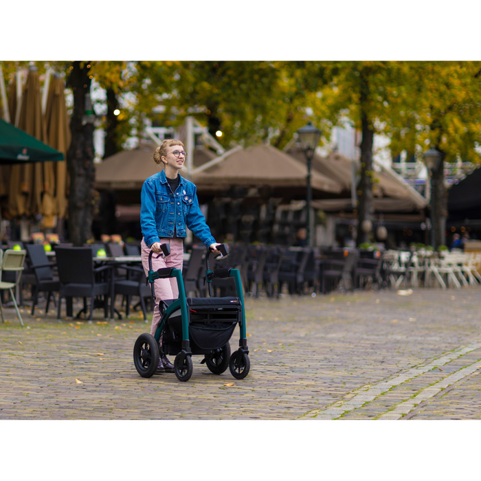 Wearing a blue jacket and pink pants, a person uses the Rollz Motion Performance All-Terrain 2-In-1 Rollator Transport Chair with rear air-filled tires to smoothly navigate the cobblestone path. Outdoor cafes with tables and umbrellas are in the background, and trees with autumn leaves line the scenic stroll.