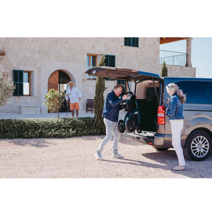 Near a stone house, a couple loads a Rollz Motion Performance All-Terrain 2-In-1 Rollator Transport Chair into a van. The woman, aided by another man in shorts and a hat, holds a cane. They smile as the sun shines brightly on this casual scene.