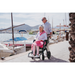 An elderly woman in a Rollz Motion Performance All-Terrain 2-In-1 Rollator Transport Chair with rear air-filled tires is pushed by an older man in sunglasses. She wears a pink outfit and hat near a marina, where the sunny setting with boats and sky suggests a cheerful outing by the water.