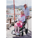 An elderly woman in a Rollz Motion Performance All-Terrain 2-In-1 Rollator Transport Chair with rear air-filled tires for smooth shock absorption holds a pink hat, while a man stands behind her by the marina. They enjoy a sunny day by the water, dressed casually for this relaxed outing.