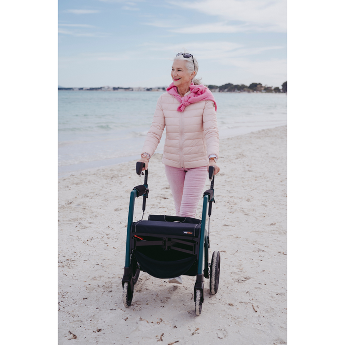 A woman with short gray hair, in a pink jacket and pants, strolls along a sandy beach using the Rollz Motion Performance All-Terrain 2-In-1 Rollator Transport Chair. Its rear air-filled tires absorb shocks well. The sea is calm and the sky is cloudy as she enjoys her peaceful walk.