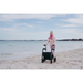 An elderly woman strolls on a sandy beach with the Rollz Motion Performance All-Terrain 2-In-1 Rollator Transport Chair, navigating smoothly with its rear air-filled tires. She wears a pink jacket and scarf as the calm sea stretches under a partially cloudy sky.